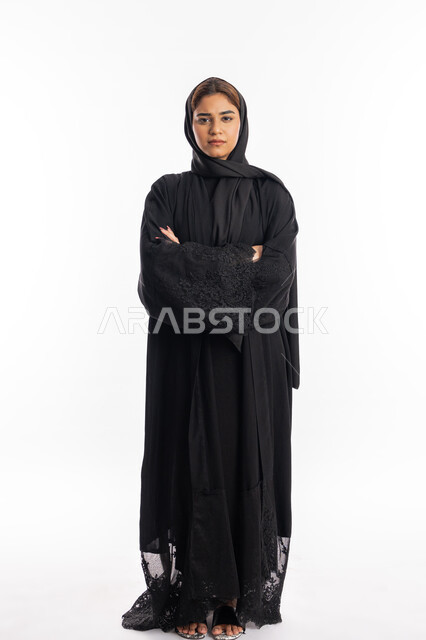 الاهتمام والعنايه بالمظهر الخارجي، شابة سعودية تقف بجدية وبأيدي متقاطعة، مفهوم الأنوثة والنعومة، بورتريه لامرأة عربية خليجية اماراتية محجبة ترتدي العباءة السوداء تنظر إلى الكاميرا بايماءات الثقة بالنفس، صورة لكامل الجسم، خلفية بيضاء