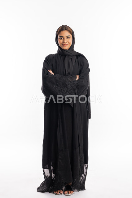 الاعتناء بالمظهر الخارجي والاهتمام بالأناقة، سيدة سعودية مبتسمة تنظر الى الكاميرا بتعابير الأنوثة والنعومة, بورتريه لإمرأة عربية خليجية إماراتية ترتدي العباءة السوداء تقف بأيدي متقاطعة, صورة الجسم بالطول الكامل, خلفية بيضاء