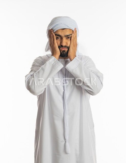 التوتر والارهاق وعدم الراحة، الاجهاد والضغط النفسي, بورتريه لشاب عربي خليجي إماراتي يرتدي العمامة والكندورة الاماراتية يضع يديه على وجهه بإيماءات التعب وعدم التصديق والذهول والانزعاج, خلفية بيضاء