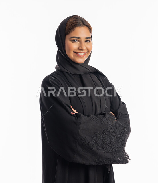 الاهتمام والعنايه بالمظهر الخارجي, أناقة النساء الخليجيات, شابة سعودية مبتسمة تقف بايدي متقاطعة، بورتريه مقرب لامرأة عربية خليجية اماراتية محجبة ترتدي عباءة سوداء تنظر إلى الكاميرا بايماءات الثقة بالنفس، خلفية بيضاء
