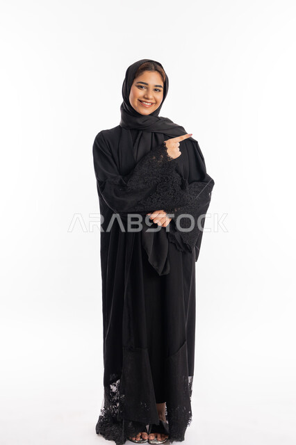 الاشارة إلى شيء ما والوقوف باستقامة، سيدة سعودية تنظر إلى الكاميرا بإيماءات السرور, تعابير التأكد و تحديد مكان معين, بورتريه لامرأة عربيه خليجيه إماراتية مبتسمة ترتدي العباءة السوداء ترفع اصبعها السبابة للاختيار، صورة الجسم بالطول الكامل, خلفية بيضاء