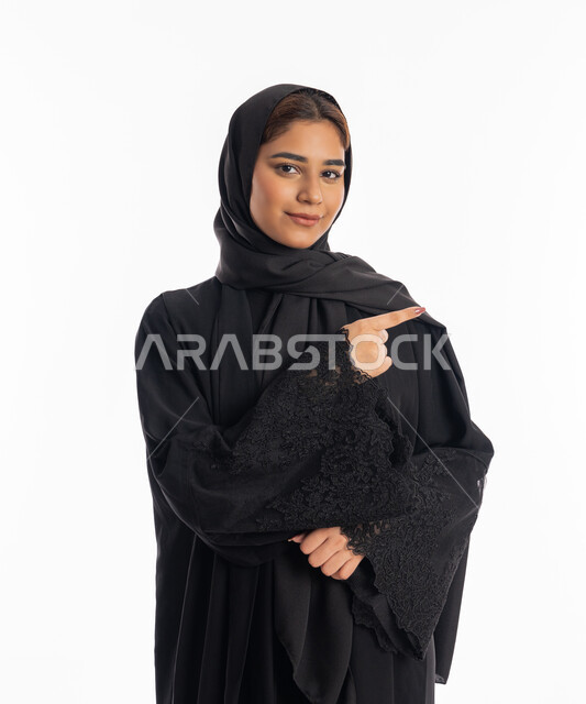 رفع إصبع السبابة لتحديد مكان معين, الوقوف باستقامة والاشارة إلى شيء ما، سيدة سعودية تنظر إلى الكاميرا بإيماءات السرور, بورتريه مقرب لامرأة عربيه خليجيه إماراتية مبتسمة ترتدي العباءة السوداء تقف بتعابير الثقة والجدية، خلفية بيضاء