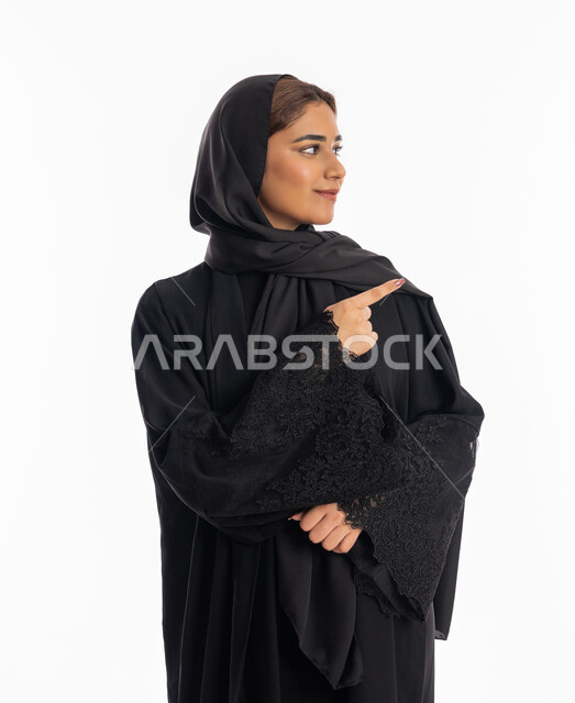 التأكد والاشارة إلى مكان معين، سيدة سعودية تنظر إلى شيء ما بإيماءات السرور, بورتريه مقرب  لامرأة عربيه خليجيه إماراتية مبتسمة ترتدي العباءة السوداء ترفع اصبعها السبابة لاختيار أمر ما, خلفية بيضاء