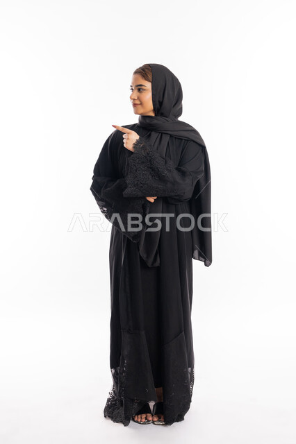 تعابير التأكد و تحديد مكان معين, سيدة سعودية تشير إلى شيء ما, بورتريه لامرأة عربيه خليجيه إماراتية مبتسمة ترتدي العباءة السوداء ترفع اصبعها السبابة للاختيار، صورة الجسم بالطول الكامل, خلفية بيضاء