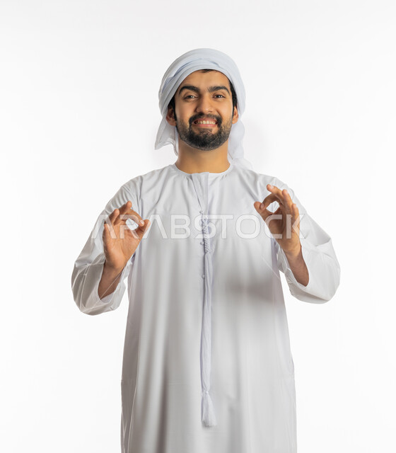 إيجاد الأفكار والحلول المناسبة, البشاشة ونظرات الحماس والشعور بالرضا, الموافقة والقبول والنصيحة، التعبير عن الجودة والاتقان، بورتريه لشاب عربي خليجي إماراتي مبتسم يرتدي الكندورة والعمامة الاماراتية يرفع يديه بإيماءات الجودة والامتياز والتميز، خلفية بيضاء