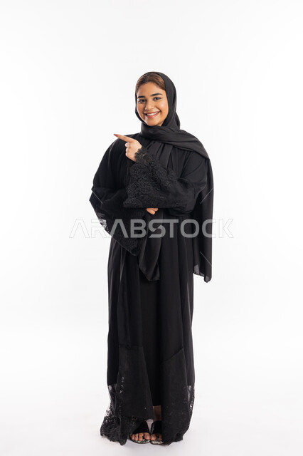 الاشارة إلى شيء ما، سيدة سعودية تنظر إلى الكاميرا بإيماءات السرور, تعابير التأكد و تحديد مكان معين, بورتريه لامرأة عربيه خليجيه إماراتية مبتسمة ترتدي العباءة السوداء ترفع اصبعها السبابة للاختيار، صورة الجسم بالطول الكامل, خلفية بيضاء