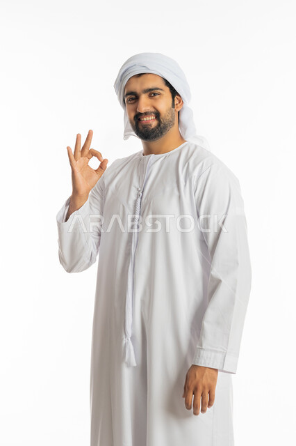 البشاشة ونظرات الحماس والشعور بالرضا, الموافقة والقبول والنصيحة، التعبير عن الجودة والاتقان، بورتريه لشاب عربي خليجي إماراتي مبتسم يرتدي الكندورة والعمامة الاماراتية يرفع يديه بإيماءات الجودة والامتياز والتميز، خلفية بيضاء