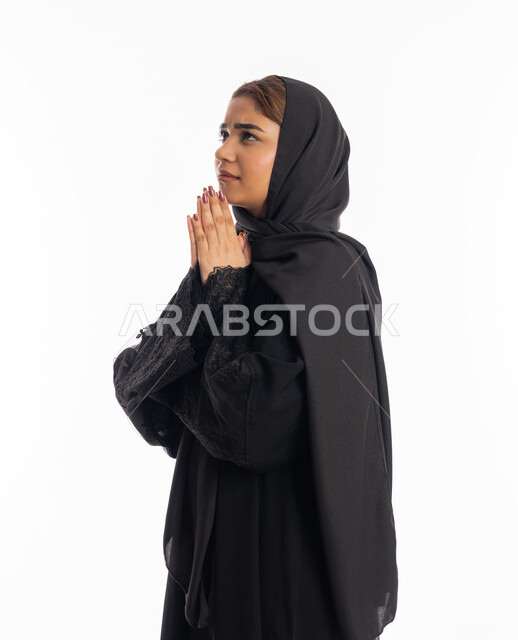 طلب العون والرغبة في تحقيق الأمنيات, الدعاء والتوسل والاسترحام, الطلب بترجي وتذلل من رب العباد, التضرع إلى الله تعالى, بورتريه من الجانب لشابة عربية خليجية اماراتية ترتدي العباءة السوداء تنظر إلى الأعلى, سيدة سعودية ترفع يديها بخشوع واستعطاف, خلفية بيضاء