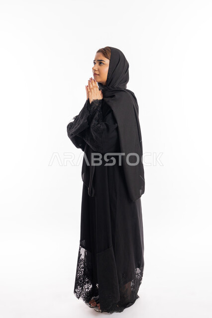 التقرب إلى الله تعالى والرغبة في تحقيق الأمنيات, الدعاء والتضرع والتوسل بإيماءات الخشوع, الترجي والتذلل لرب العالمين, سيدة سعودية ترتدي العباءة السوداء تنظر إلى الأعلى, بورتريه لإمرأة عربية خليجية إماراتية ترفع يديها باستعطاف, الجسم بالطول الكامل, خلفية بيضاء