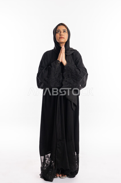 الدعاء والتضرع والتوسل بإيماءات الخشوع, التقرب إلى الله تعالى والرغبة في تحقيق الأمنيات, الترجي والتذلل لرب العالمين, سيدة سعودية ترتدي العباءة السوداء تنظر إلى الأعلى, بورتريه لإمرأة عربية خليجية إماراتية ترفع يديها باستعطاف, الجسم بالطول الكامل, خلفية بيضاء