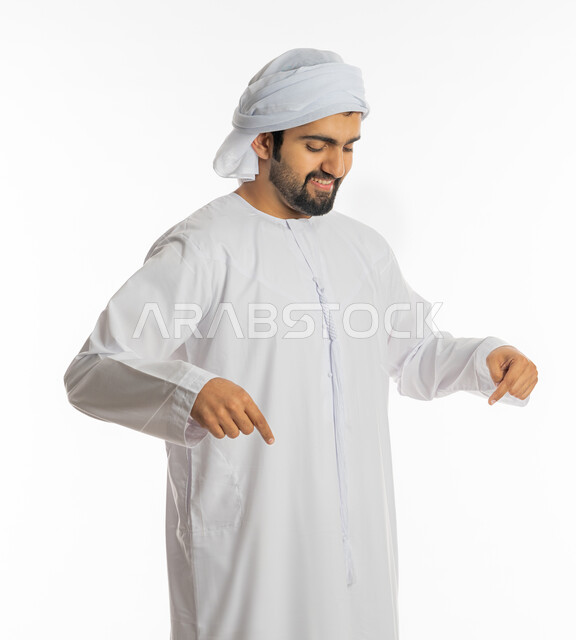 اشارات تحديد هذا المكان (هنا)، حركات وتعابير وجود شيء ما، تعين موقع محدد، بورتريه لرجل عربي خليجي اماراتي مبتسم يرتدي العمامة والكندورة يشير بأصابعه نحو الأسفل, خلفية بيضاء