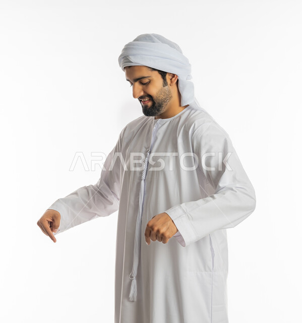 حركات وتعابير وجود شيء ما، اشارات تدل على هذا المكان (هنا)، تحديد موقع معين، بورتريه لرجل عربي خليجي اماراتي مبتسم يرتدي العمامة والكندورة يشير بأصابعه نحو الأسفل, خلفية بيضاء