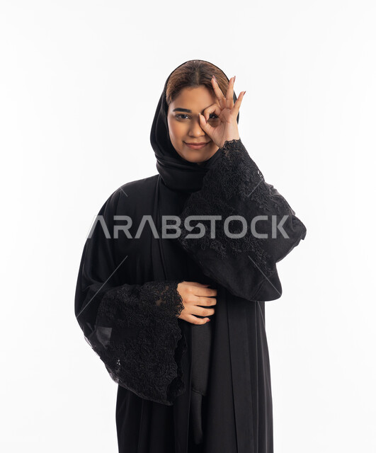 الوقوف والنظر إلى الكاميرا, حب البحث والاستكشاف والمغامرة, مفهوم الرؤية المستقبلية، بورتريه لامرأة عربية خليجية إماراتية محجبة مبتسمة ترتدي عباءة سوداء تحاول تقريب الأشياء البعيدة, سيدة سعودية تضع يديها على عينها بشكل عدسة, خلفية بيضاء