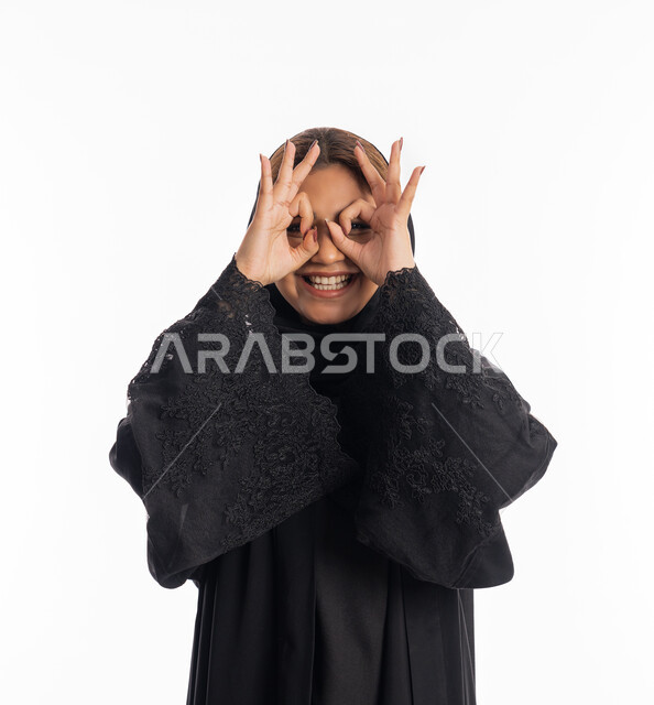 حب البحث والاستكشاف والمغامرة, مفهوم الرؤية المستقبلية، بورتريه مقرب لامرأة عربية خليجية إماراتية محجبة مبتسمة ترتدي عباءة سوداء تحاول تقريب الأشياء البعيدة, سيدة سعودية تضع يديها على عينها بشكل عدسة, خلفية بيضاء