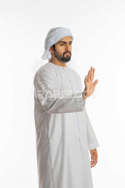 الوقوف بتهديد والنهي عن فعل شيء ما, الصمت والتوقف عن الكلام, بورتريه لرجل عربي خليجي اماراتي يرتدي الكندورة والعمامة الاماراتية يرفع كفه للأعلى بإيماءات الرفض وعدم الرضى والقبول, خلفية بيضاء