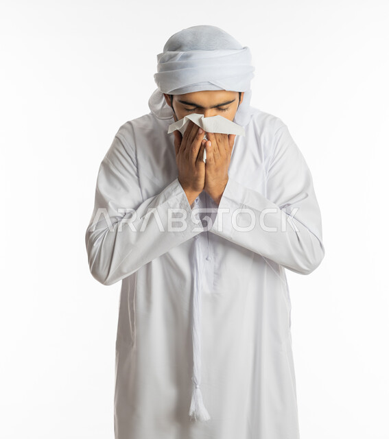 مفهوم الاهتمام بالصحة والحاجة إلى العلاج والدواء, استخدام المناديل الورقيه للعطس, الشعور بالمرض و الزكام والتهاب الأنف التحسسي, بورتريه لشاب عربي اماراتي خليجي يرتدي العمامة والكندورة يضع يديه على وجهه بإيماءات العطاس, خلفية بيضاء