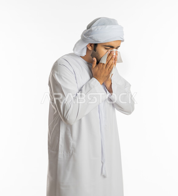 الشعور بالمرض و الزكام والحاجة الى العلاج والدواء, التهاب الأنف التحسسية, استخدام المناديل الورقيه للعطس, بورتريه لشاب عربي اماراتي خليجي يرتدي العمامة والكندورة يضع يديه على وجهه بإيماءات العطاس, خلفية بيضاء