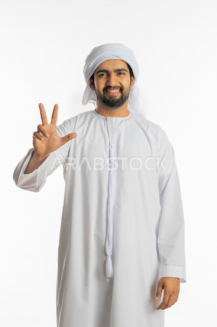 الفوز والنصر والحب, علامات وإشارات مشعة بالأمل والتفاؤل, الوقوف باستقامة والنظر إلى الكاميرا, بورتريه لشاب عربي خليجي إماراتي يرتدي الكندورة والعمامة يرفع يده بعلامة ثلاثية, المثابرة والتميز والتحديات لتحقيق النجاح, خلفية بيضاء