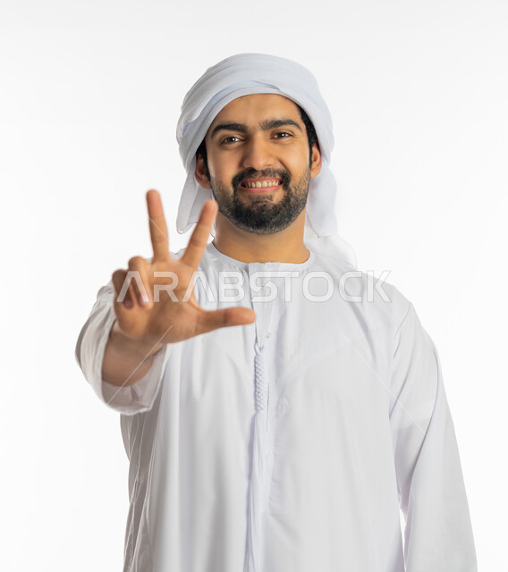علامات وإشارات مشعة بالأمل والتفاؤل, الوقوف باستقامة والنظر إلى الكاميرا, بورتريه لشاب عربي خليجي إماراتي يرتدي الكندورة والعمامة يرفع يده بعلامة ثلاثية, الفوز والنصر والحب, المثابرة والتميز والتحديات لتحقيق النجاح, خلفية بيضاء