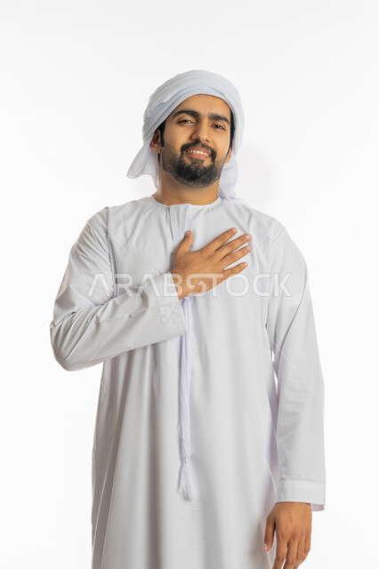 الاحساس بمشاعر رومنسية مليئة بالسعادة, الوقوع في الغرام والحب, بورتريه لشاب عربي خليجي إماراتي يرتدي الكندورة والعمامة يضع يديه على صدره بإيماءات العشق, تعابير وحركات شبابية, خلفية بيضاء