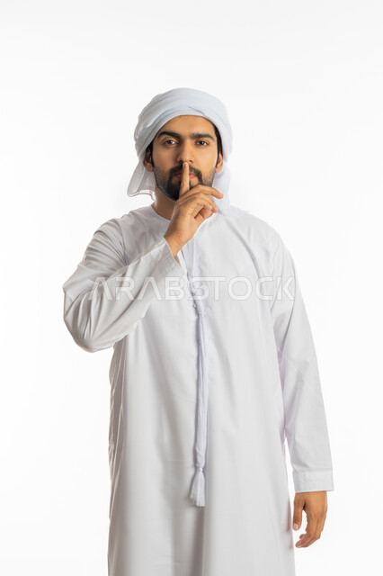 الوقوف بجدية والنظر إلى الكاميرا, التوقف عن الكلام وخفض الصوت, الهدوء والسكون وعدم الإزعاج، بورتريه لرجل عربي خليجي إماراتي يرتدي الكندورة الاماراتية والعمامة يضع أصبع السبابة على فمه بإيماءات عدم التدخل وطلب السكوت والصمت, خلفية بيضاء