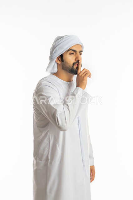 التوقف عن الكلام وخفض الصوت, الهدوء والسكون وعدم الإزعاج، بورتريه لرجل عربي خليجي إماراتي يرتدي الكندورة الاماراتية والعمامة يضع أصبع السبابة على فمه بإيماءات عدم التدخل وطلب السكوت والصمت, خلفية بيضاء