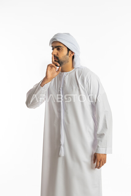 Calm, quiet, and not disturbed, a portrait of an Emirati Gulf Arab man wearing the Emirati kandora and turban, placing his index finger on his mouth with gestures of non-interference and asking for silence and silence, to stop talking and lower the volume, white background.