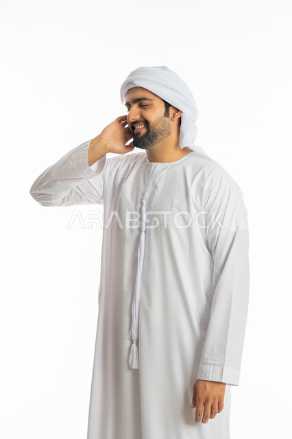 الاستمتاع بالغناء وسماع الموسيقى بتعابير التفاعل والاندماج, التسلية والترفيه وقضاء الوقت الممتع, مفهوم الاصغاء والاستماع, بورتريه لشاب عربي خليجي اماراتي يرتدي الكندورة والعمامة يضع يده على إذنه بإيماءات الطرب والسعادة, خلفية بيضاء