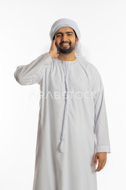 التسلية والترفيه وقضاء الوقت الممتع, مفهوم الاصغاء والاستماع, بورتريه لشاب عربي خليجي اماراتي يرتدي الكندورة والعمامة يضع يده على إذنه بإيماءات الطرب والسعادة, الاستمتاع بالغناء وسماع الموسيقى بتعابير التفاعل والاندماج, خلفية بيضاء