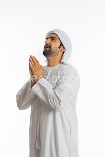 Standing with gestures of supplication, supplication, and supplication, asking with hope and humility from the Lord of servants, supplicating to God Almighty, a portrait of an Arab Gulf Emirati young man wearing a turban and an Emirati kandora, raising his hands in humility and entreaty, asking for help and the desire to fulfill wishes, white background.