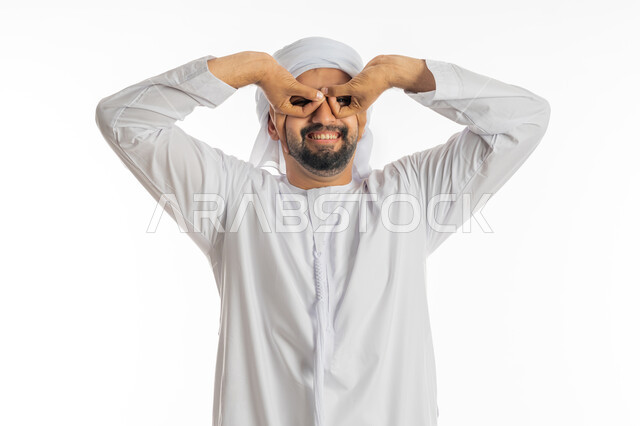 Love of discovery, knowledge and adventure, joy and pleasure in seeing something, trying to bring distant things closer, planning future goals and ambitions, close-up portrait of a smiling Emirati Gulf Arab man wearing a turban and kandora, placing his hands over his eyes, white background.