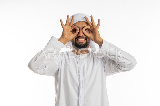 البشاشة والسرور برؤية شيء ما, محاولة تقريب الأشياء البعيدة, التخطيط للأهداف والطموحات المستقبلية, بورتريه مقرب لشاب عربي خليجي إماراتي مبتسم يرتدي العمامة والكندورة يضع يديه على عينيه, حب الاكتشاف والمعرفة والمغامرة, خلفية بيضاء