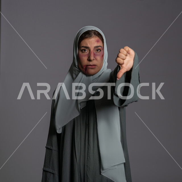 آثار الكدمات الجسدية, 25 نوفمبر اليوم العالمي للقضاء على العنف ضد النساء ,حقوق المرأة المسلمه, العلاقات الزوجية الفاشلة, عدم القبول و التقييم السلبي, بورتريه مقرب لشابة عربيه إماراتية خليجية محجبة تشير بإصبع الابهام نحو الاسفل بتعابير عدم الرضا, سيدة سعودية حزينة, خلفية رمادية