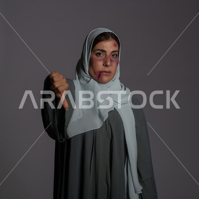 عدم القبول و التقييم السلبي, آثار الكدمات الجسدية, 25 نوفمبر اليوم العالمي للقضاء على العنف ضد النساء ,حقوق المرأة المسلمه, العلاقات الزوجية الفاشلة, بورتريه مقرب لشابة عربيه إماراتية خليجية محجبة تشير بإصبع الابهام نحو الاسفل بتعابير عدم الرضا, سيدة سعودية حزينة, خلفية رمادية