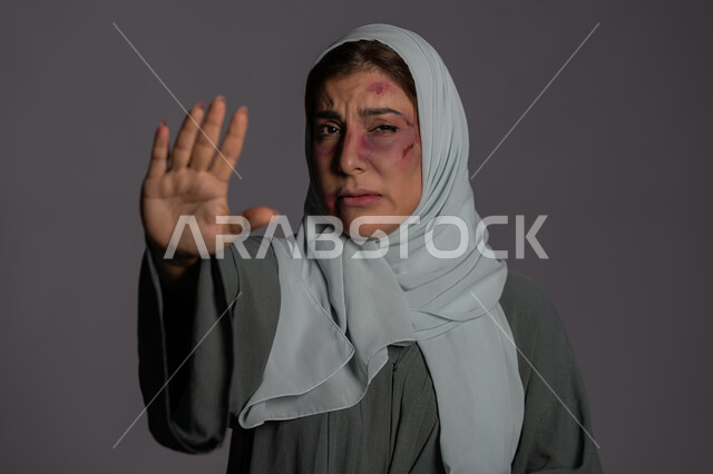 Expressions of crying and fatigue, failed marital relationships, November 25, International Day for the Elimination of Violence against Women, Muslim women’s rights, physical effects of bruises, a close-up portrait of a veiled Arab Emirati Gulf woman raising her palm up with gestures asking to stop, a sad Saudi woman, gray background