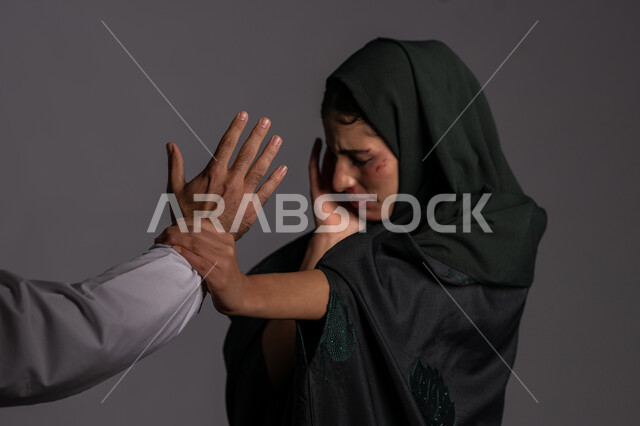 Stop harming Muslim women, a man’s palm raised with a hitting gesture, marital problems and psychological illnesses, November 25, International Day for the Elimination of Violence against Women, the effects of physical bruises, a close-up portrait of a veiled Emirati Gulf Arab young woman trying to defend herself, a sad Saudi woman, background grey