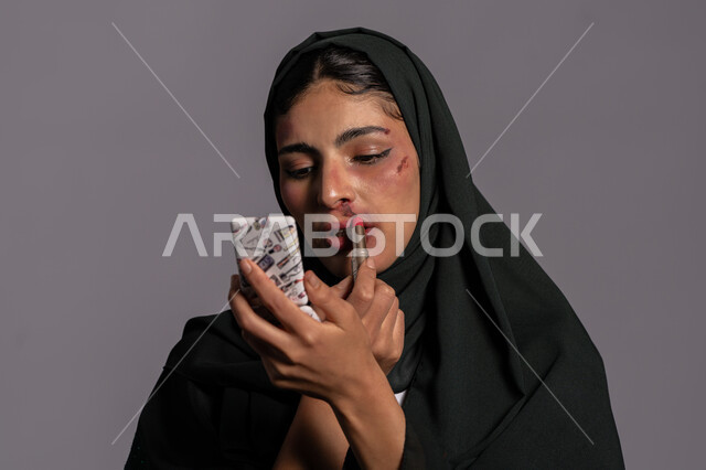 November 25, International Day for the Elimination of Violence against Women, psychological illnesses and marital problems, trying to hide the effects of physical bruises, a close-up portrait of a veiled Emirati Gulf Arab young woman holding a mirror and a red pen in her hand, a sad Saudi woman, Muslim women’s rights, gray background