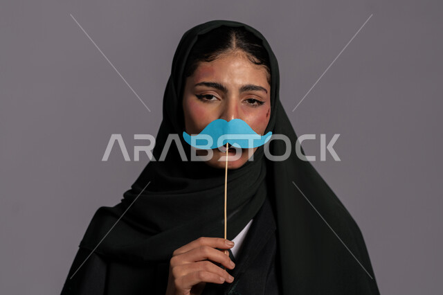 آثار الكدمات الجسدية, بورتريه مقرب لشابة عربيه إماراتية خليجية محجبة تحمل بيدها قصاصة ورقية على شكل شارب, 25 نوفمبر اليوم العالمي للقضاء على العنف ضد النساء ,سيدة سعودية حزينة, اجبار الامرأة على السكوت من قبل الرجال, حقوق المرأة المسلمه، امراض نفسية ومشاكل زوجية, خلفية رمادية