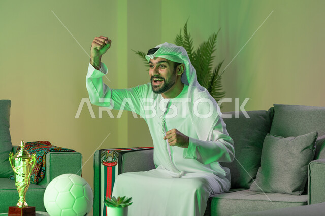Enjoyment of watching the national team and the competition, a young Arab Gulf Emirati man wearing the kandura and white ghutra and raising his hands with gestures of enthusiasm, integration, interaction and sports encouragement, expressions of happiness with victory and victory, spending enjoyable times watching the match