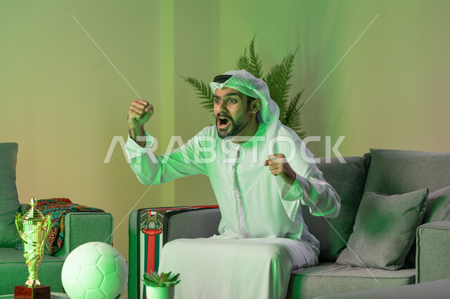 تعابير النصر والفوز، الاندماج والتفاعل والتشجيع الرياضي، العاب المباريات الترفيهية، ايماءات السعادة بالربح والانتصار، شاب عربي خليجي اماراتي يرتدي الكندورة والغترة البيضاء، يرفع يديه بإيماءات الحماس، الاستجمام وقضاء أوقات ممتعة بمشاهدة المباراة
