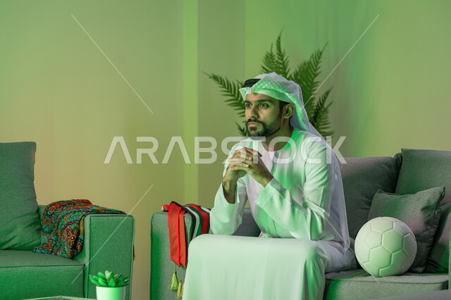 العاب المباريات الترفيهية، شاب عربي خليجي اماراتي يرتدي الكندورة والغترة البيضاء، الجلوس على الاريكة وبجانبه كرة، شبك الأيدي ببعضها بإيماءات التوتر والتفكير بشيء ما، الاندماج في مشاهدة المباراة والفريق المفضل