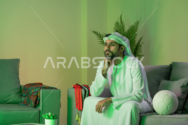 انشطة ترفيهية مسلية، قضاء أوقات ممتعة بمشاهدة المباراة، الاندماج في تشجيع الفريق الرياضي، شاب عربي خليجي اماراتي يرتدي الكندورة والغترة البيضاء و يضع يده على وجهه، الجلوس على الاريكة وبجانبه كرة، إيماءات التأمل والتفكير بشيء ما