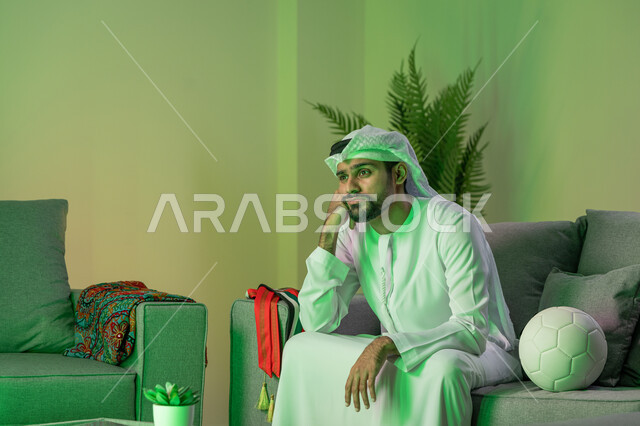 قضاء أوقات ممتعة بمشاهدة المباراة، شاب عربي خليجي اماراتي يرتدي الكندورة والغترة البيضاء و يضع يده على وجهه، الجلوس على الاريكة وبجانبه كرة، إيماءات الحزن و الخذلان لخسارة الفريق المفضل