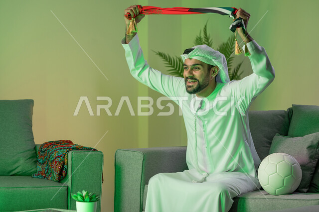 Enjoying watching the national team, spending free time watching the matches, integration, interaction and sports encouragement, a young Arab Gulf Emirati man wearing the kandora and white ghutra, raising his hands and carrying the scarf of the team he supports, sitting on the sofa with a ball next to him, gestures of happiness with victory and triumph
