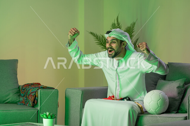 قضاء وقت الفراغ بمشاهدة المباريات، الاندماج والتفاعل والتشجيع الرياضي، نشاط منزلي ترفيهي فردي، شاب عربي خليجي اماراتي يرتدي الكندورة والغترة البيضاء و يرفع يديه بإيماءات الحماس والسرور، الجلوس على الاريكة وبجانبه كرة، تعابير السعادة بالفوز والانتصار