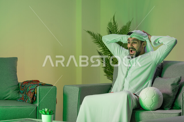 Following and watching matches, an individual home entertainment activity, an Arab Gulf Emirati young man wearing a kandura and a white ghutra, putting his hands behind his head and a smile on his face, sitting on the sofa with a ball next to him, expressions of happiness with victory and victory, integration, interaction and sports encouragement