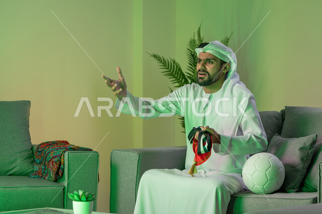 الشغف بمشاهدة ومتابعة مباراة كرة القدم, الاندماج والتفاعل والتشجيع الرياضي, شاب عربي خليجي اماراتي يرتدي الكندورة والغترة البيضاء و يحمل بيده وشاح فريقه المفضل، الجلوس على الاريكة وبجانبه كرة، إيماءات الغضب والانزعاج