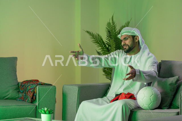 الاندماج والتفاعل والتشجيع الرياضي, الشغف بمشاهدة ومتابعة مباراة كرة القدم, شاب عربي خليجي اماراتي يرتدي الكندورة والغترة البيضاء و يرفع يديه بإيماءات الغضب، الجلوس على الاريكة وبجانبه كرة، تعابير الحزن لخسارة الفريق المفضل