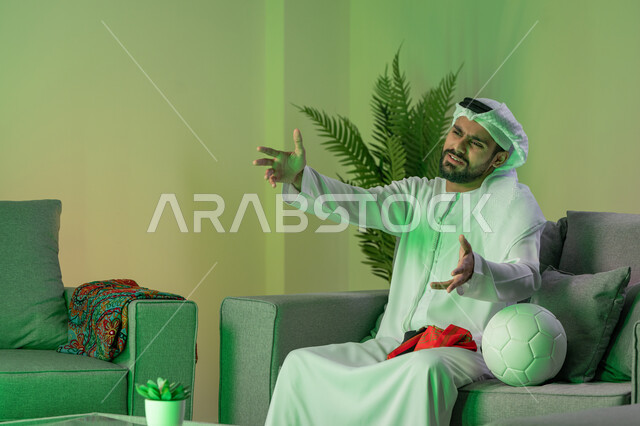 ايماءات الحزن لخسارة الفريق المفضل، شاب عربي خليجي اماراتي يرتدي الكندورة والغترة البيضاء و يرفع يديه بإيماءات الانزعاج، الجلوس على الاريكة وبجانبه كرة، مشاهدة الالعاب الرياضية