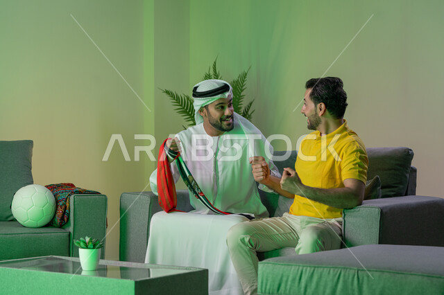 مشاهدة المباريات وتشجيع الفريق، ايماءات الحماس بالفوز وتسجيل الأهداف، الشغف بمتابعة المباريات، قضاء وقت ممتع مع الأصدقاء في المنزل، صديقان عربيان خليجيان اماراتيان يشاهدان مباراة كرة القدم في غرفة المعيشة